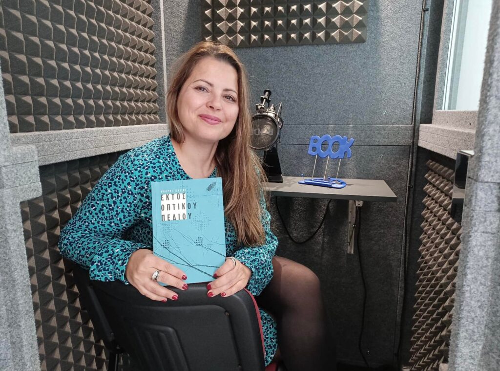 Η Αργυρώ Σπυριδάκη κάθεται στο στούντιο ηχογράφησης του Διαβάζω για τους Άλλους και κρατάει το βιβλίο με τίτλο: Εκτός Οπτικού Πεδίου. 